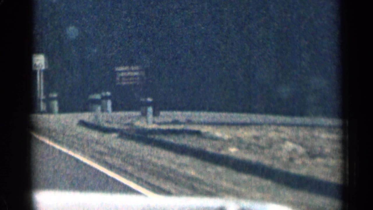 1963 Colorado moving-car frame showing a two-lane road shoulder, short roadside posts, a small distant sign, and dark mountain hillside in faded 8mm color.