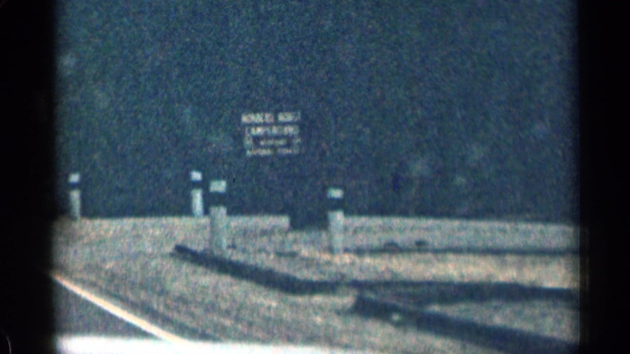 Soft roadside view with a sign beyond a paved edge and forested background.