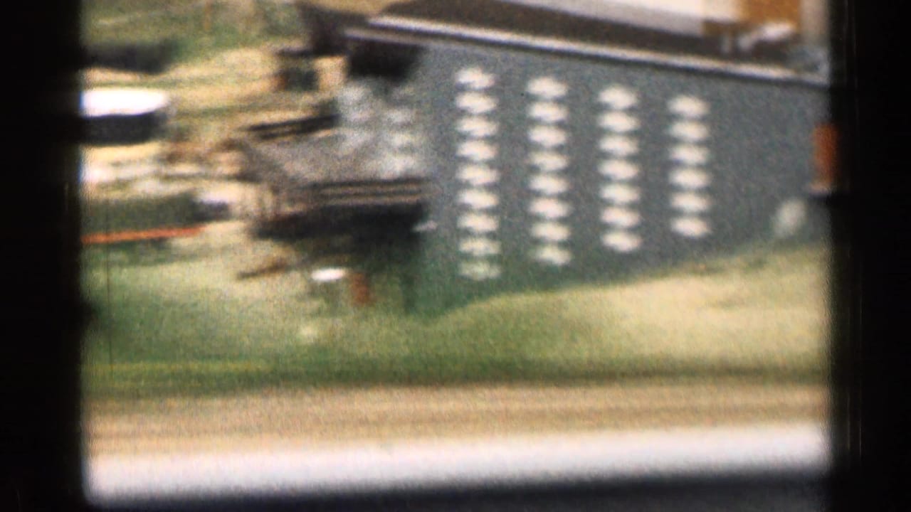 Motion-blurred 8mm frame from a moving vehicle showing a parking area, cars, a low building, and repeated light markings on a wall or sign.