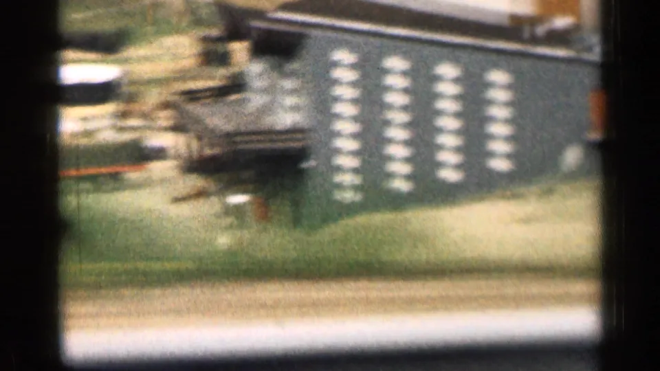 Motion-blurred 8mm frame from a moving vehicle showing a parking area, cars, a low building, and repeated light markings on a wall or sign.