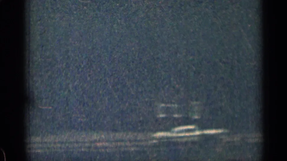 Low-light 8mm frame showing faint horizontal lights and dark reflective water near the end of the night drive clip.