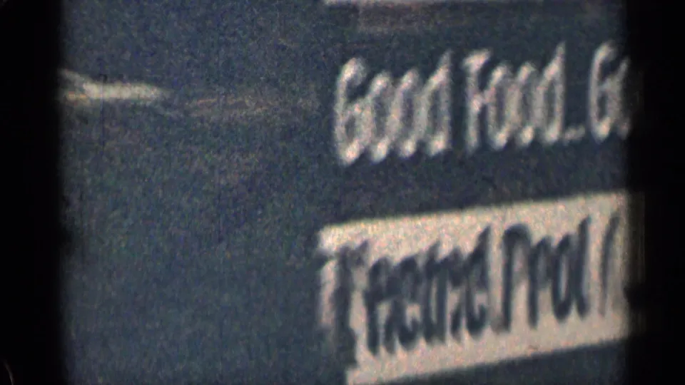 Blurred roadside motel or lodge sign in 1963 Colorado footage, showing Good Food and Overnight Guest lettering.