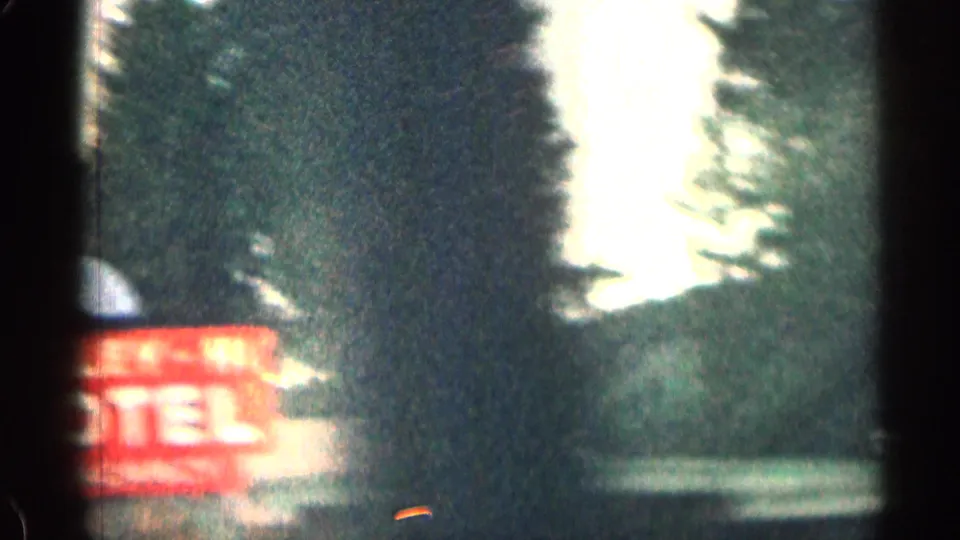 1963 Colorado car-window frame showing a red motel sign beside evergreen trees and a bright gap in the road scene.