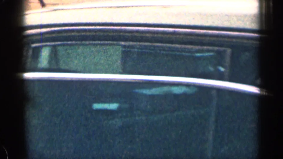 A grainy 1963 home-movie frame shows part of a dark car or vehicle window area with reflective trim and soft focus.