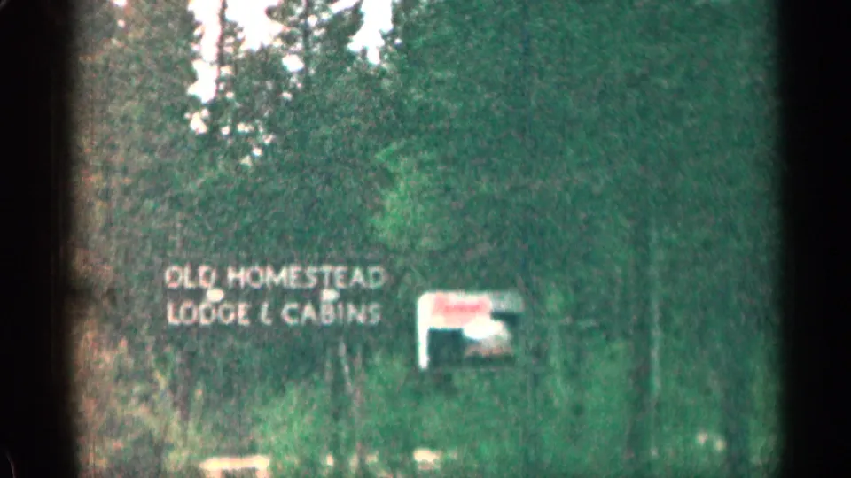 1963 Colorado home-movie frame showing the Old Homestead Lodge & Cabins sign near brush, trees, and a smaller roadside sign.