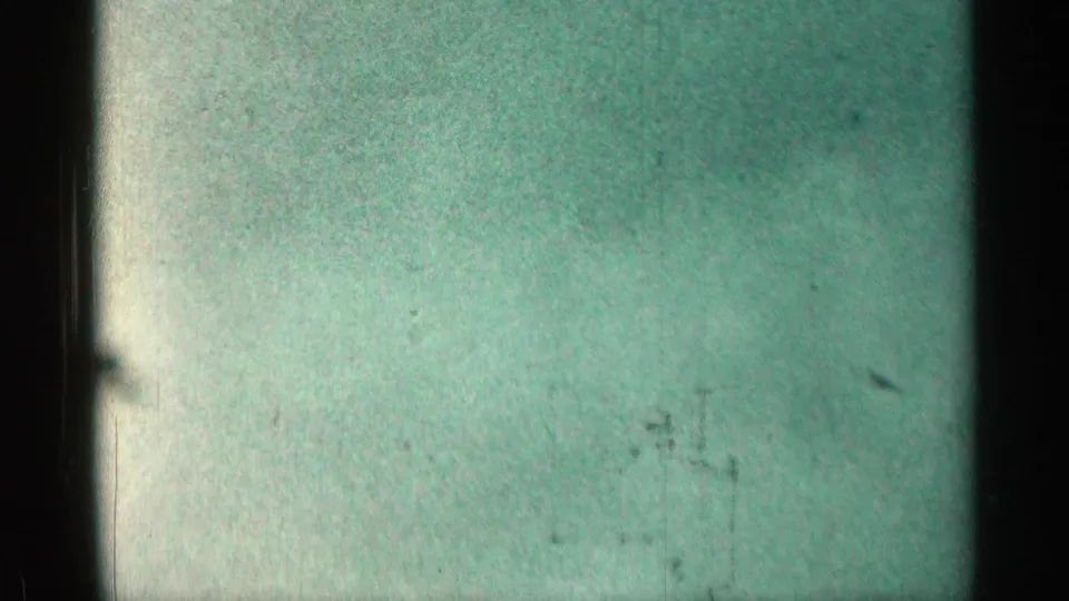 Greenish 8mm frame with little visible scene detail, showing grain, scratches, exposure variation, and dark film edges.