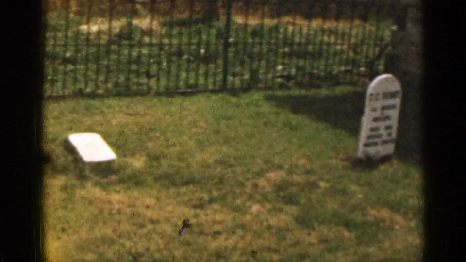 White cemetery headstone in a grassy area beside a black iron fence, filmed on 8mm in Colorado.