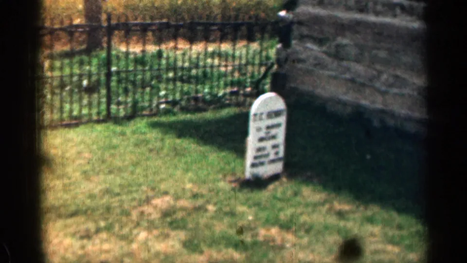 White headstone beside a black iron fence and stone wall in a small cemetery setting in Colorado.