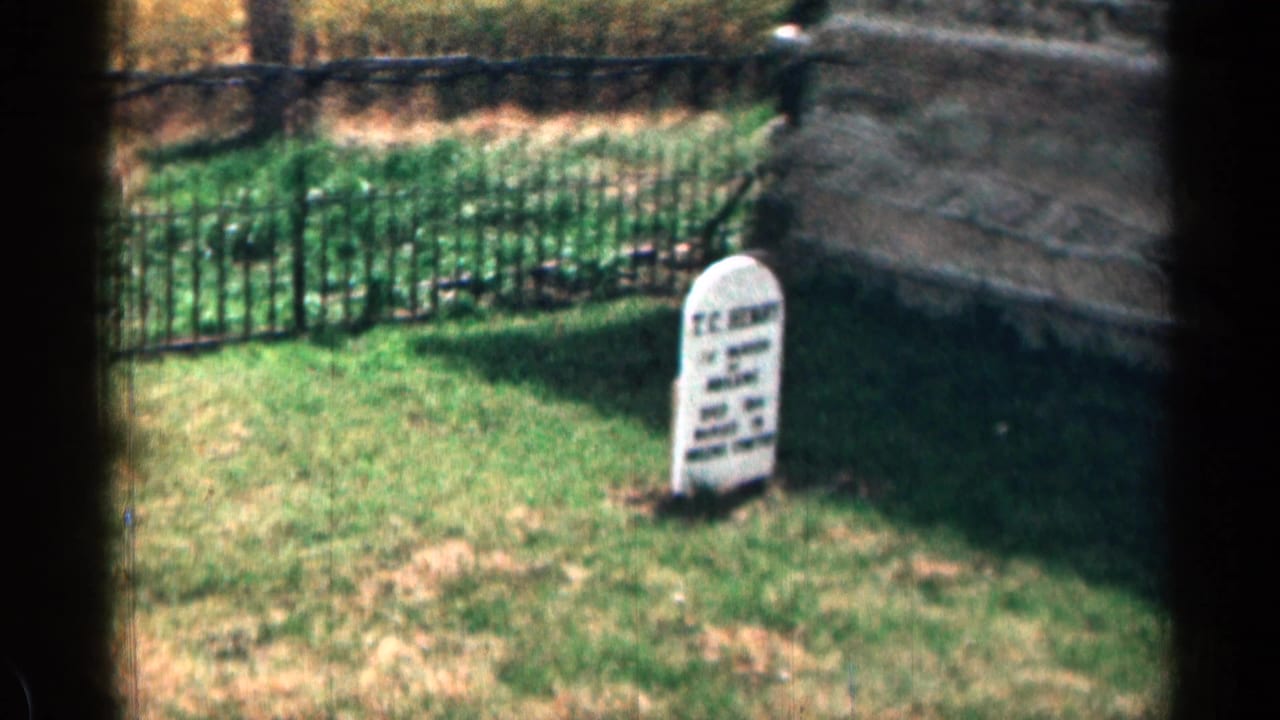 1963 8mm Colorado cemetery view with a white headstone in sunlit grass, an iron fence behind it, and stonework at right.