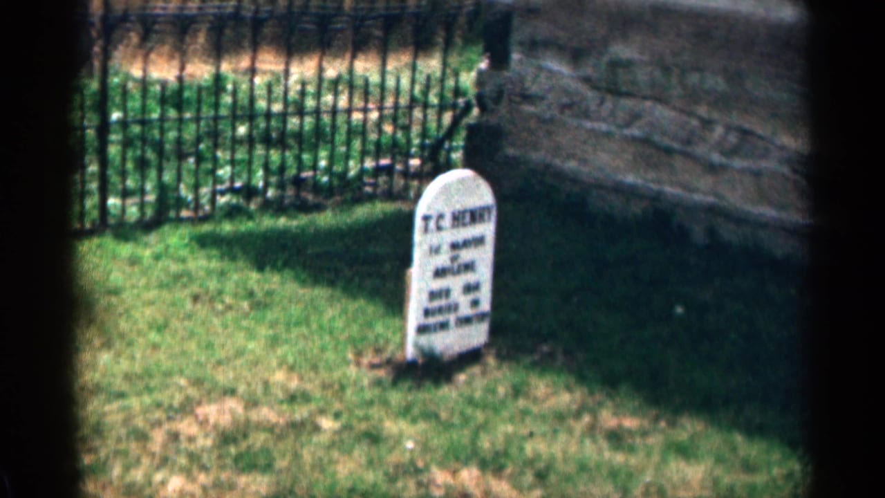 White T.C. Henry headstone beside grass, iron fence, and stonework.