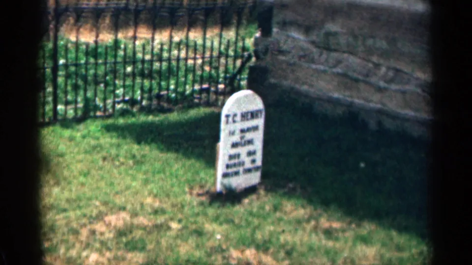 1963 8mm cemetery frame showing a white T.C. Henry headstone in a grassy plot with a black iron fence behind it.