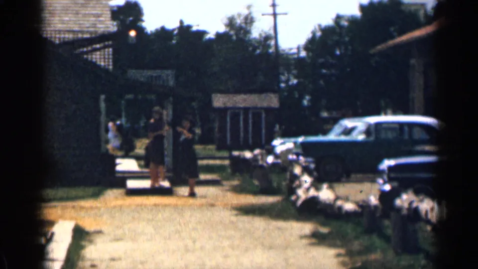 Colorado 1963 home-movie frame of two distant women, parked 1960s cars, a sidewalk, houses, and trees.