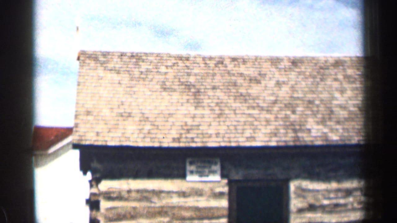 1963 Colorado 8mm frame showing a small log cabin exterior with a shingled roof, dark doorway, and pale sky behind it.