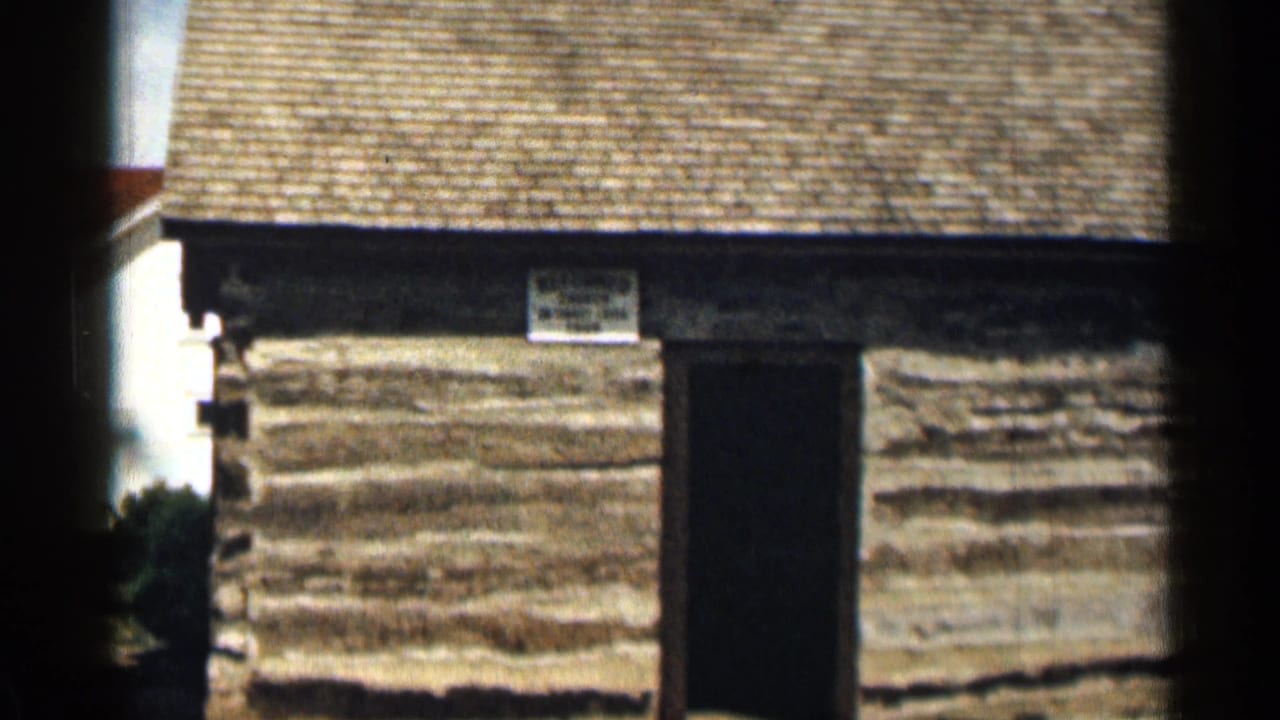 1963 Colorado 8mm frame with the cabin centered, showing stacked log walls, a dark entrance, roof shingles, and a small sign above the door.
