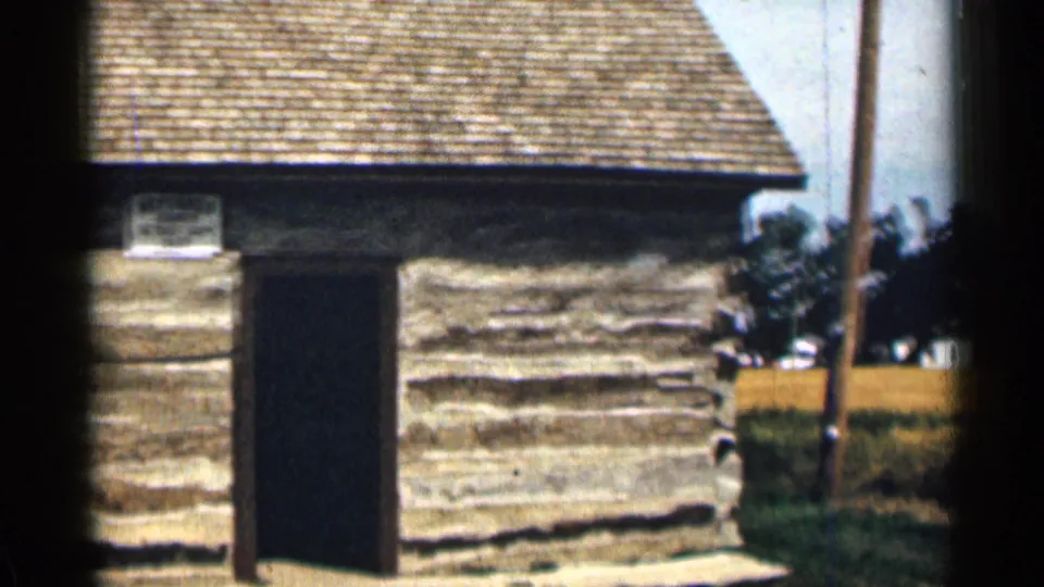 1963 Colorado 8mm frame showing the cabin entrance, rough log walls, small sign, grass foreground, and a utility pole at the side.