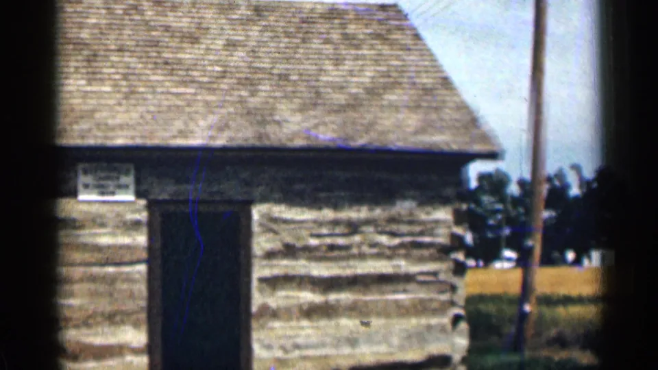 1963 Colorado 8mm frame showing the log cabin exterior with the doorway, roof shingles, trees, utility pole, and a low fence or barrier nearby.
