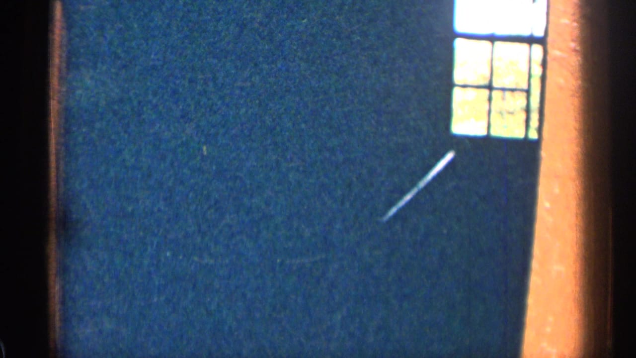 1963 Colorado home-movie frame with a small daylight window, dark wall, orange trim, and a pale diagonal streak.