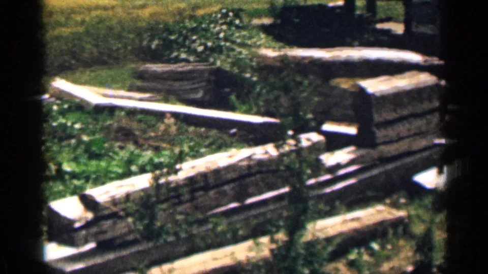 1963 Colorado home-movie frame showing a closer yard view with white planks, stone blocks, grass, and surrounding greenery.