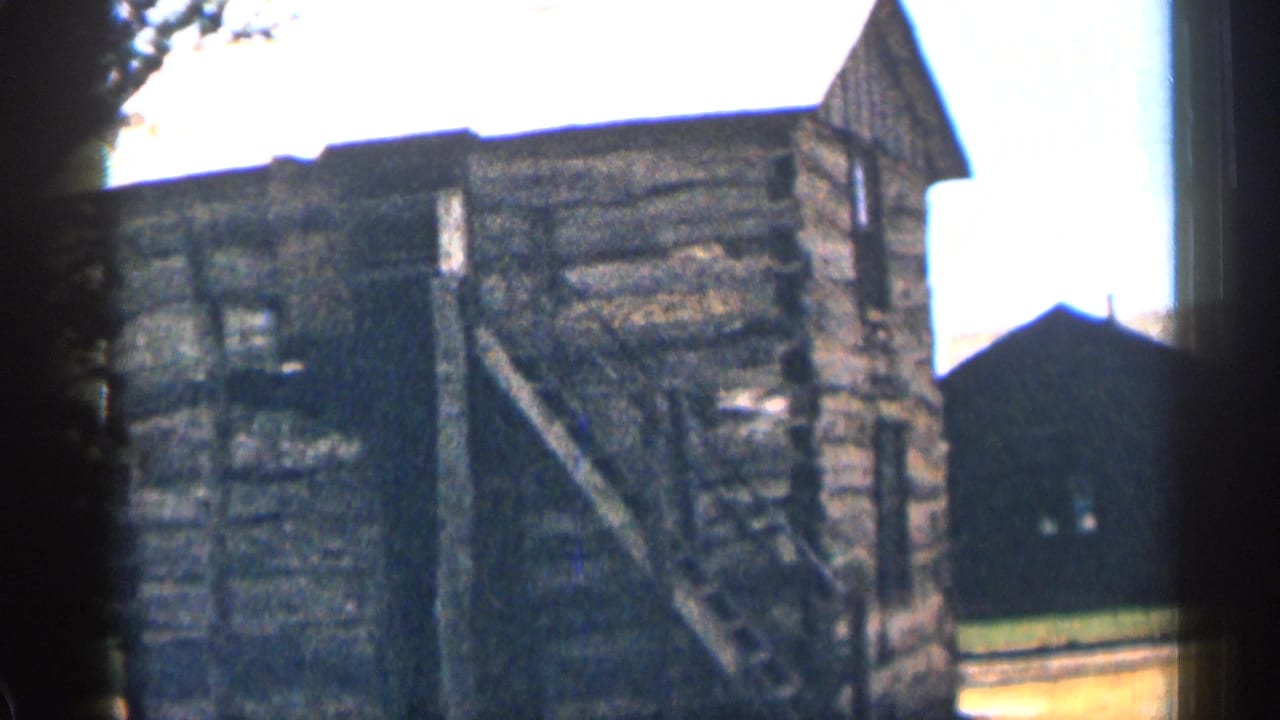 Log cabin with sloped roof, exterior stair, grass, and dark outbuilding behind it.