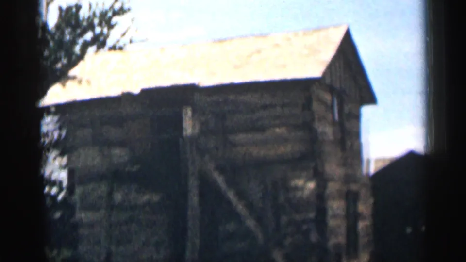 1963 Colorado home-movie frame with a closer view of the log cabin roofline, rough timber walls, exterior stair, and tree edge.