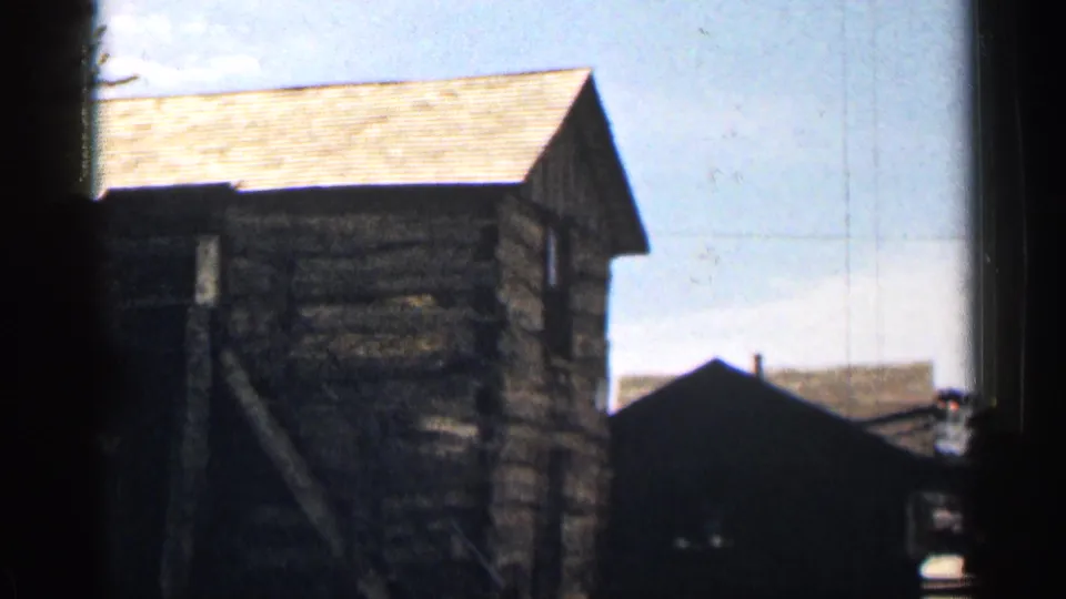 1963 Colorado frame showing the side of a rustic log cabin with shingled roof, exterior stair, open sky, and a neighboring dark building.