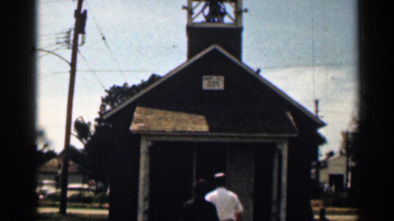 1963 Colorado 8mm frame showing a small dark schoolhouse with bell tower, porch roof, sign, and two people walking toward the doorway.