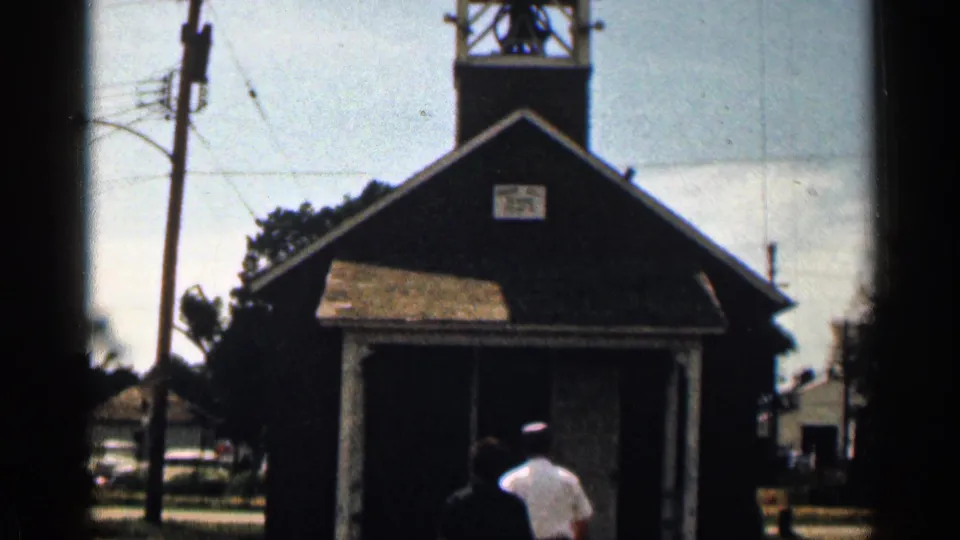 1963 Colorado 8mm frame showing a small dark schoolhouse with bell tower, porch roof, sign, and two people walking toward the doorway.