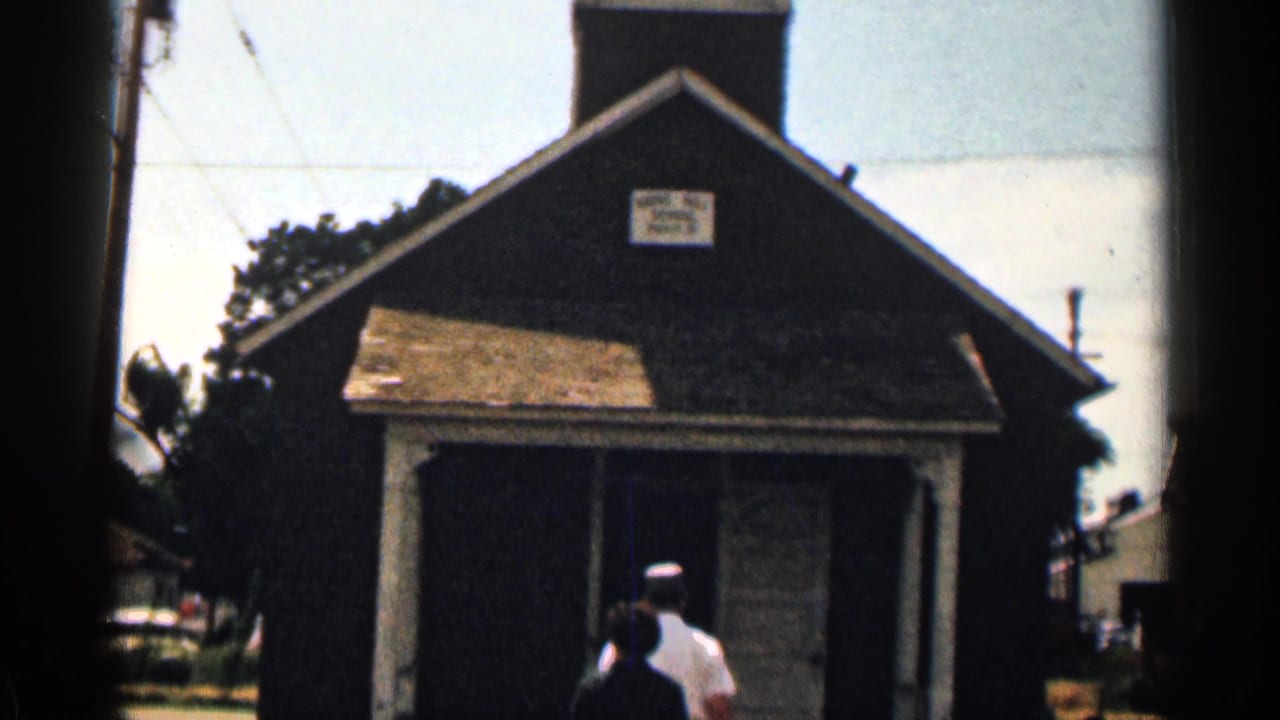 A small Colorado schoolhouse facade appears in 8mm footage, with a readable sign above the entrance and two figures moving inside.