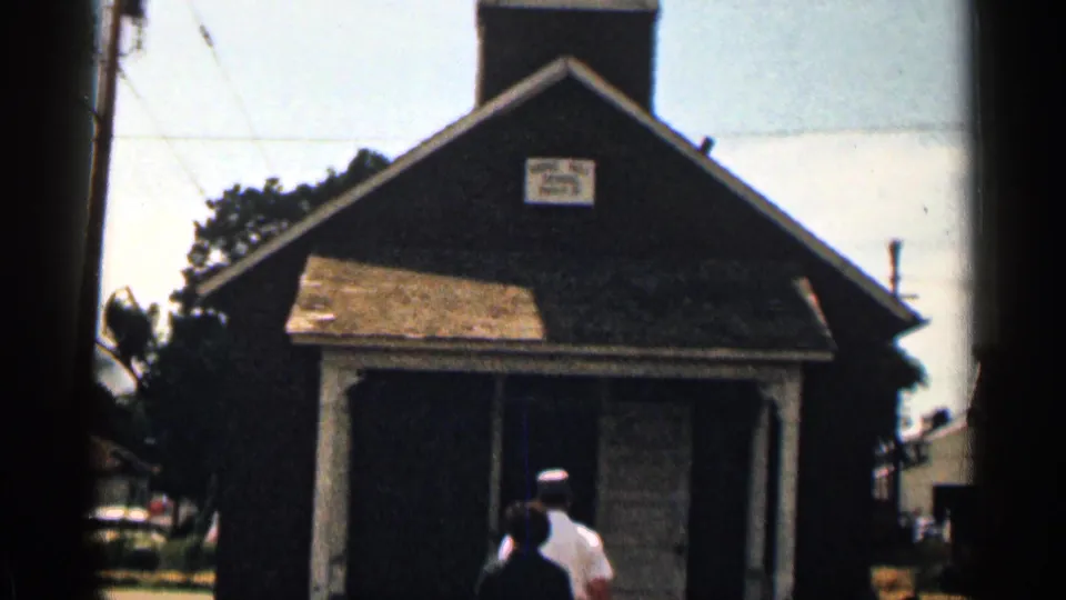 A small Colorado schoolhouse facade appears in 8mm footage, with a readable sign above the entrance and two figures moving inside.