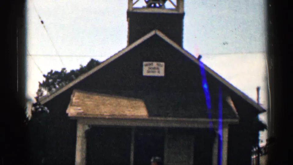 Handheld 8mm view of the Colorado schoolhouse front, showing the bell tower, gabled roof, porch roof, entrance, and school district sign.