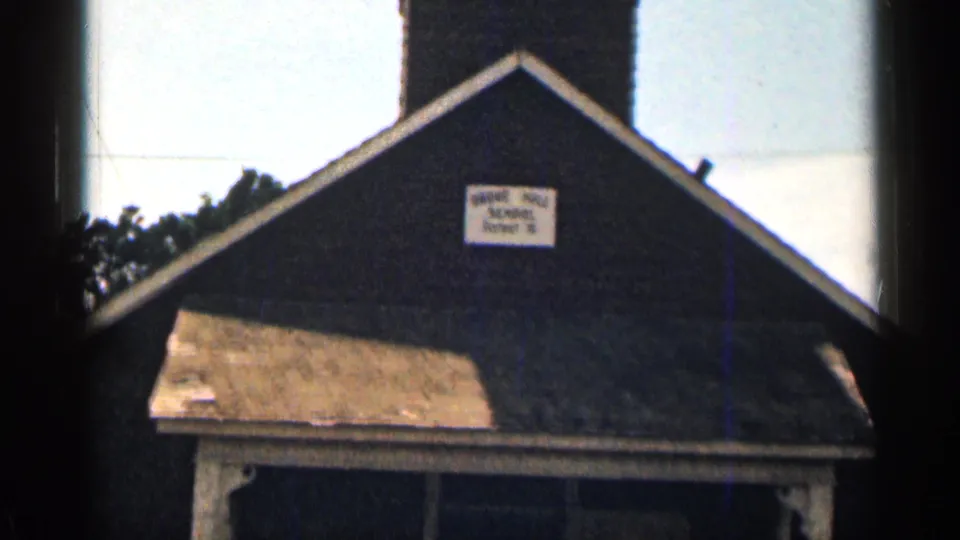 Close 1963 Colorado frame of the small schoolhouse exterior, with the school sign, dark facade, porch roof, and entrance filling most of the view.