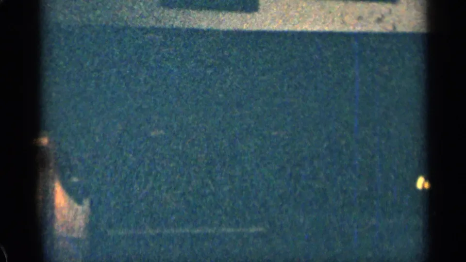 Dark interior-looking frame from the 1963 8mm clip, with bluish wall tones, heavy grain, scratches, and partial edge detail.