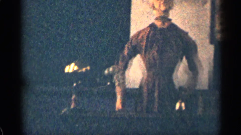 A mannequin-like figure stands in a dim interior, its pale torso and dark garment visible against a shadowed wall.