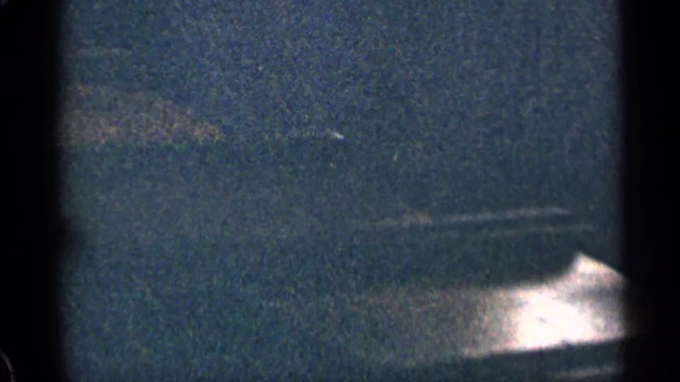 Soft, grainy 8mm frame showing a dark exterior or pavement-like area with a bright sunlit patch near the bottom edge.