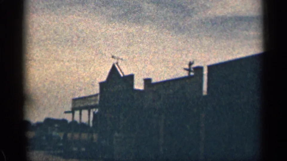 1963 Colorado home-movie frame looking down a quiet street between dark building fronts, with porch lines, roof shapes, and utility poles visible.
