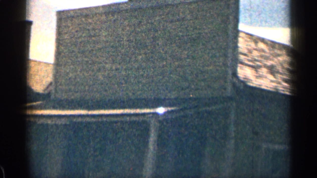1963 Colorado 8mm frame showing a dark weathered building exterior, roof edge, pale sky, and heavy film grain.