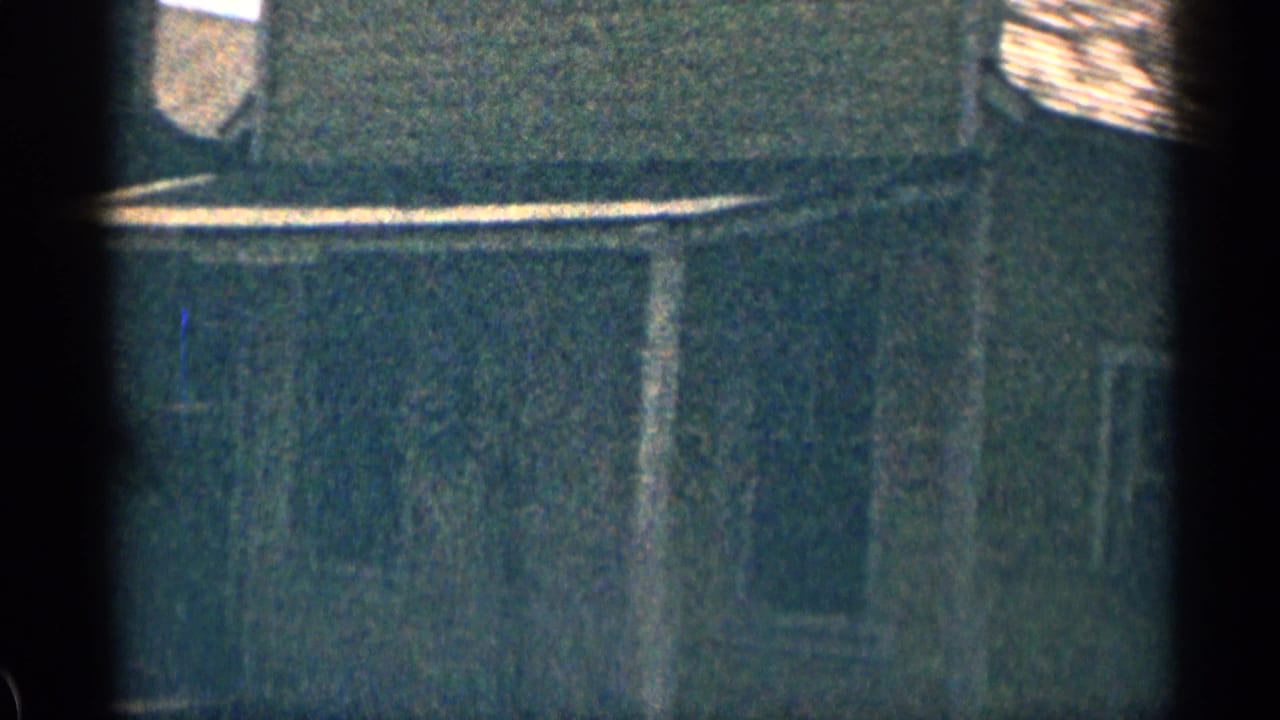 1963 Colorado home-movie frame of a weathered exterior wall, low roofline, and dark vertical film-frame obstruction.