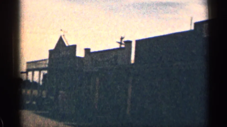 1963 Colorado 8mm frame showing silhouetted weathered buildings, porch posts, rooftops, and bright daylight sky.