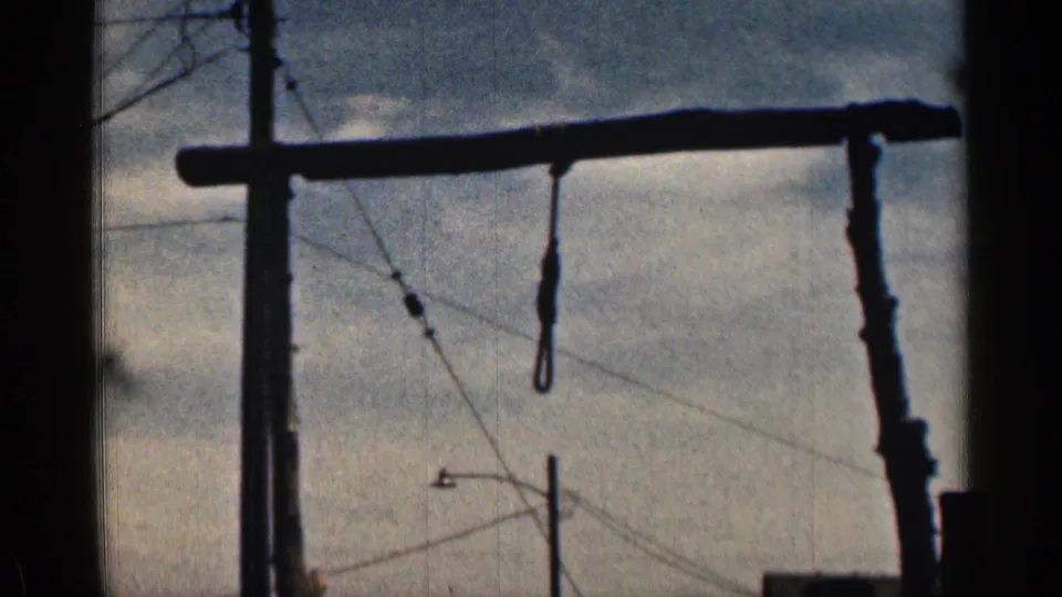 1963 Colorado frame showing the gallows crossbeam and noose silhouetted against cloudy sky, with utility poles and wires in the background.