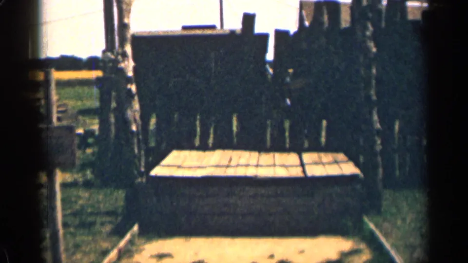 1963 Colorado 8mm frame showing a plank platform beneath the gallows, with dark boards, grass, and bright open background.