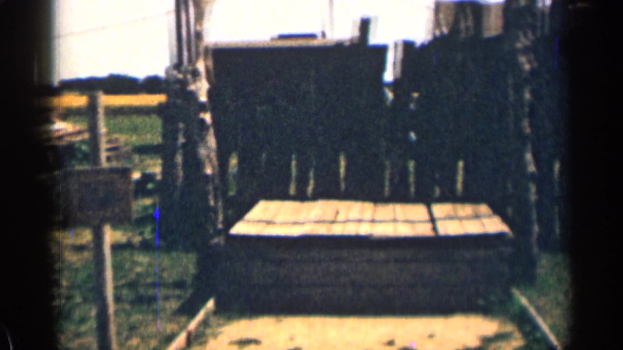 1963 Colorado home-movie frame with a low wooden structure, dark fence boards, grass, posts, and partial roofline in the background.