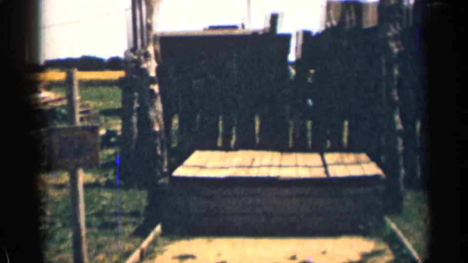 1963 Colorado home-movie frame with a low wooden structure, dark fence boards, grass, posts, and partial roofline in the background.