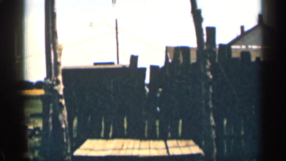 1963 Colorado 8mm frame with dark rough fencing, a low wooden platform, posts, grass, and overexposed sky.