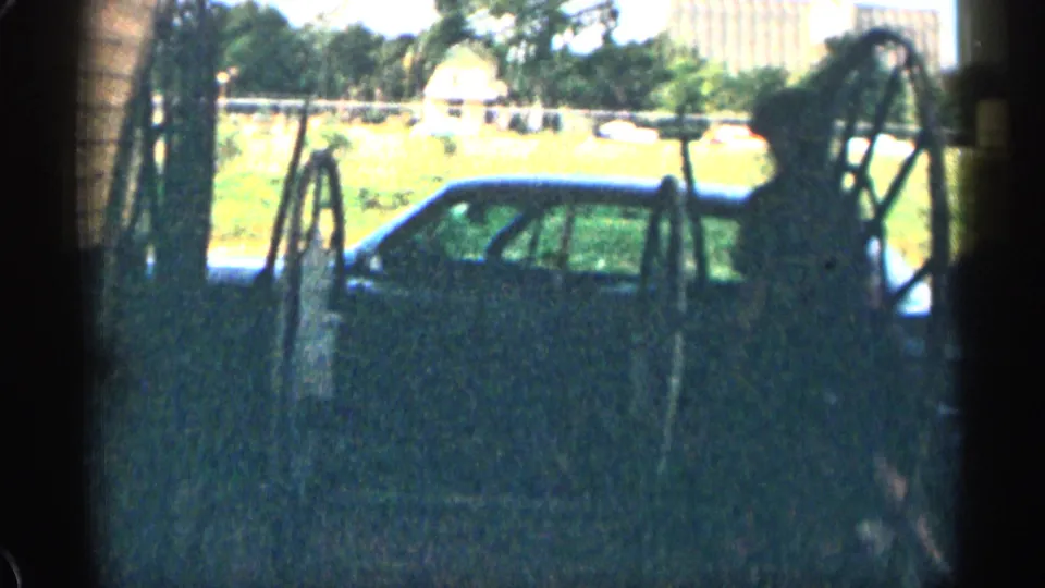 1963 Colorado home-movie frame with a vintage car parked beside fencing and rustic structures, with bright open ground behind it.