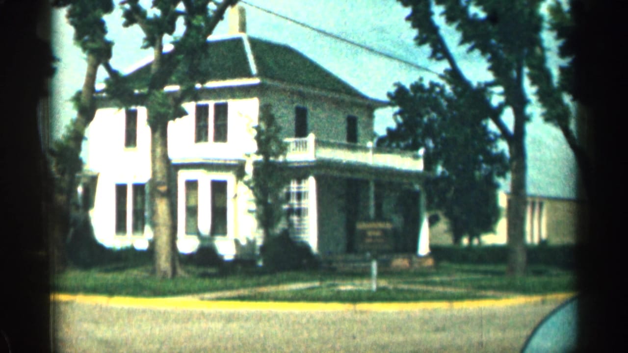 1963 Colorado street view of a white two-story house with a wraparound porch, trees, lawn, and yellow curb.
