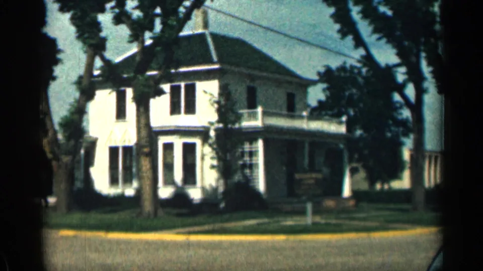 1963 Colorado 8mm frame of a white house across the street, with porch railings, tree shadows, lawn, and yellow curb.