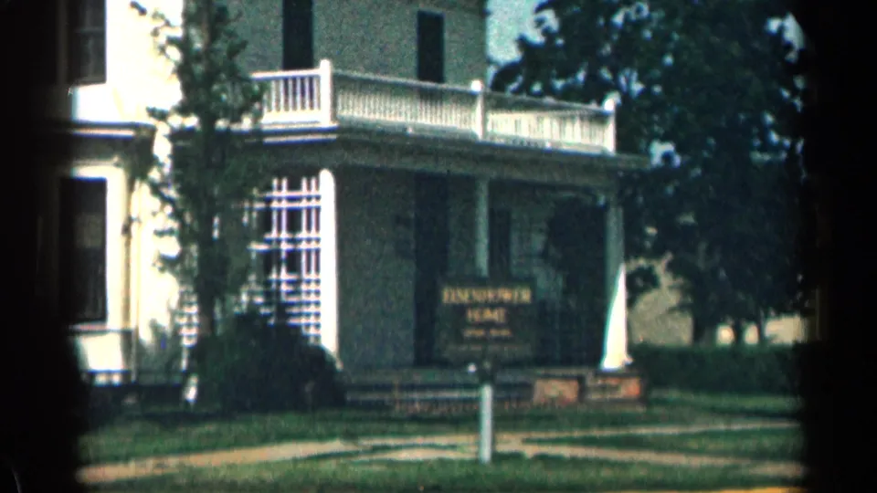 Closer 1963 Colorado 8mm frame of the Eisenhower Home porch area, with balcony railing, lattice window, sign, lawn, and sidewalk visible.