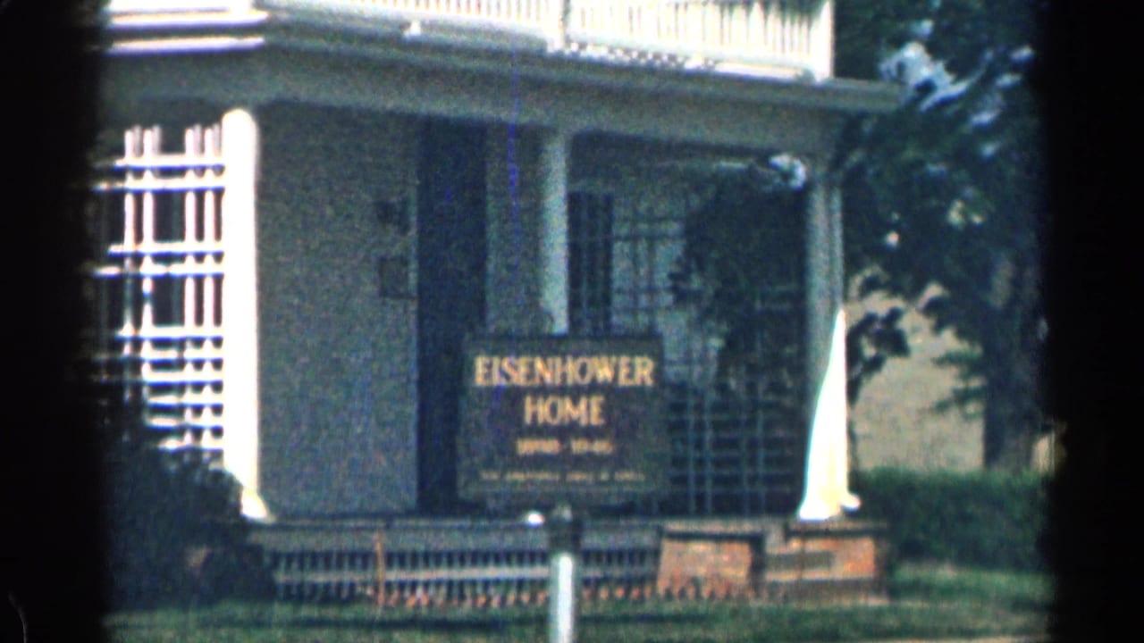 Second 1963 Colorado frame of the Eisenhower Home sign, porch columns, dark doorway, window grilles, lawn, and shrubs.