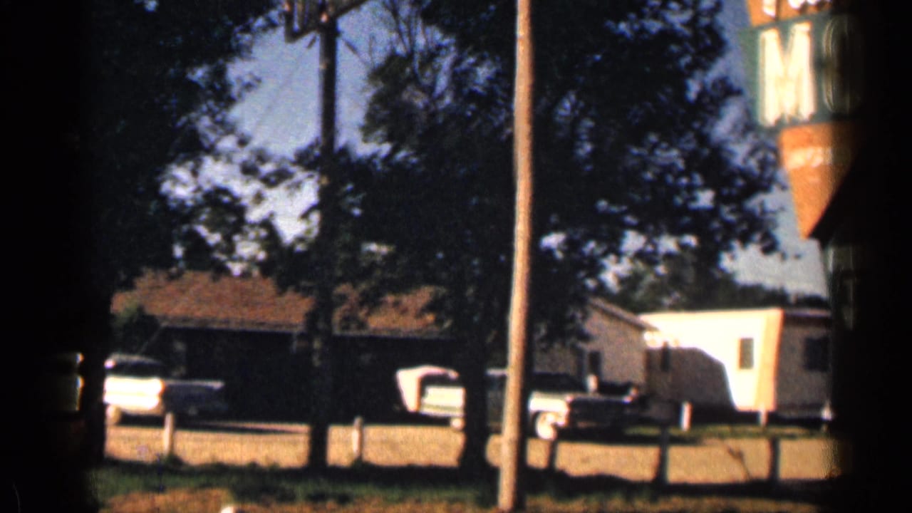 1963 Colorado roadside motel view with a pickup, trailer, trees, utility poles, and low buildings in bright daylight.