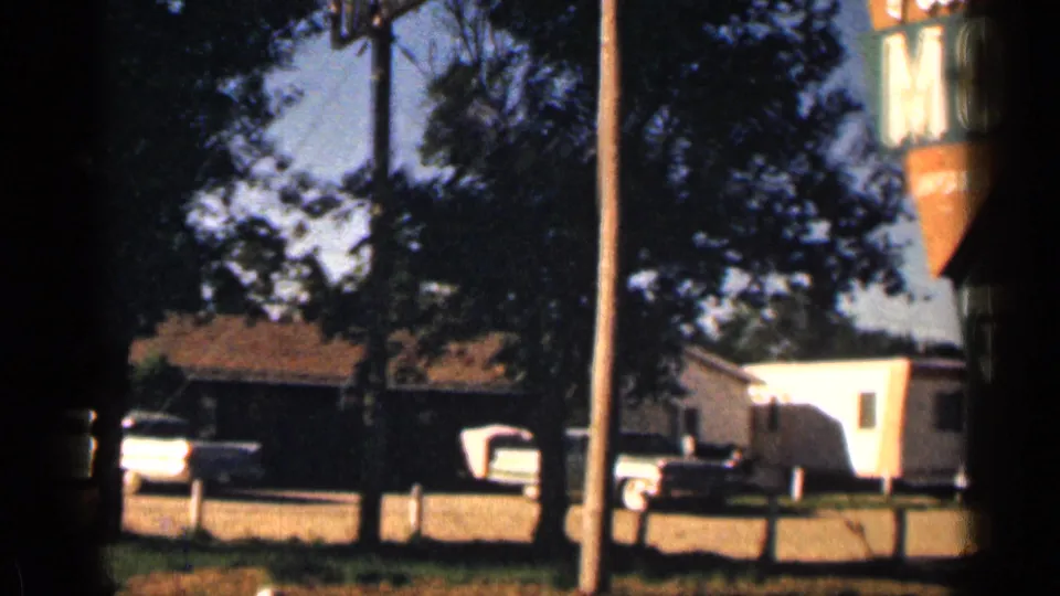 1963 Colorado roadside motel view with a pickup, trailer, trees, utility poles, and low buildings in bright daylight.